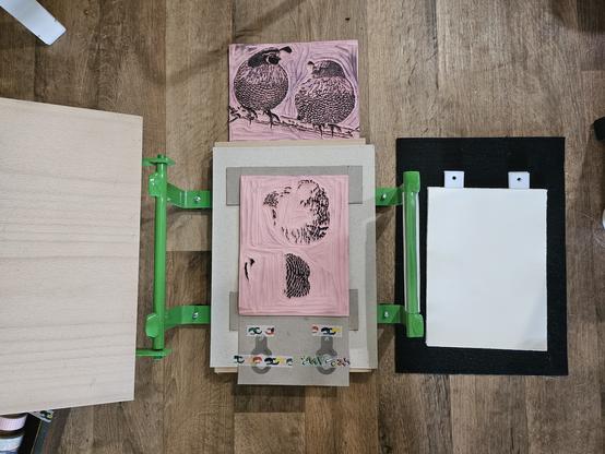 an open blockprinting press. sitting on the press is a carved block in a registration jig made of scrap cardboard, with registration pins taped down. the second block is on the floor next to it; so is a pile of papers with registration tabs washi taped to them