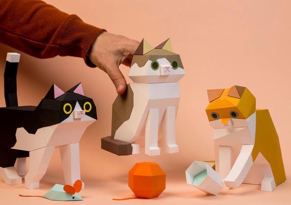 A person places a paper cat model among two other paper cat models on a surface. A toy mouse and a toy ball are nearby.