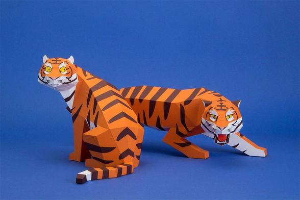 Two paper craft tigers with orange and black stripes positioned against a blue background; one seated, the other crouching with mouth open.