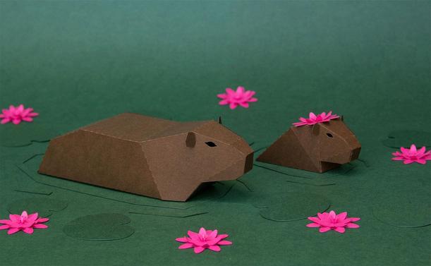 Two paper capybaras with pink flowers on their heads are placed on a green surface, surrounded by pink paper flowers.