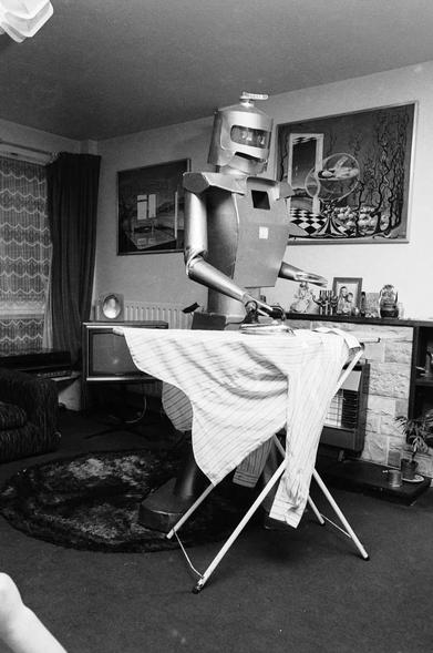 Photography. A black and white photo of a 1.80 m (6-foot 3-inch) tall robot ironing laundry. The tall silver tin man has a somewhat angular build, an oval helmet with a visor for a head, a narrow waist and arms with gripping tongs. He is standing behind the ironing board in his inventor's living room, ironing a shirt. The living room is still furnished in the typical style of the 1970s, with abstract art above the fireplace, a television and plush carpeting.
Info: The large roboter (Super lightweight at 6 1/2 pounds), was invented by Peter Holland. "Mr Robotham the Great" is a 6 foot tall walking, talking robot who can also shake hands, bend knees to sit, and all functions are operated by radio control, but only for three hours - then its batteries have to be recharged.
(Photos via Getty Images)
Note.: There is an original construction drawing on the net.