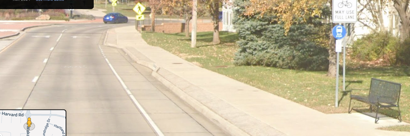 a sign with a bike symbol "may use full lane", two lanes each way going into a roundabout, which used to be stop signs with a side street, the sign is cowering behind a "bus stop" which is a bench and a stick on the curb-tight sidewalk.  A painted bike lane has a sharp ramp up to the sidewalk just before the roundabout, crosswalk.
