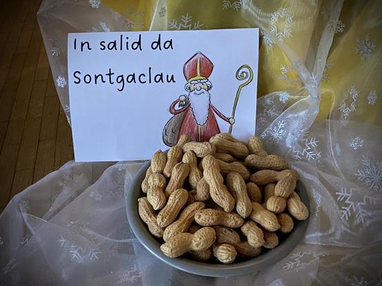 Eine Schüssel Erdnüsse ist im Bild zentriert, begleitet von einem Schild mit einer karikaturhaften Weihnachtsmannfigur und dem Text "In salid da Sotngaclau". Der Hintergrund besteht aus einem strukturierten, hellen Stoff mit Schneeflockenmustern.