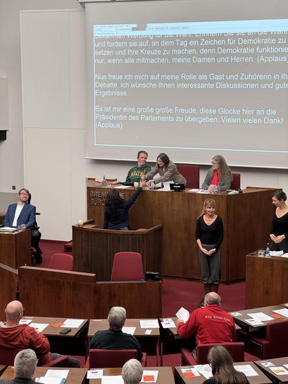 Die Vizepräsidentin der Bremischen Bürgerschaft, Sahhanim Görgü-Philipp, übergibt die Glocke zum Einläuten der Sitzung an die Präsidentin des Behinderenparlaments, Victoria Ruppnow.