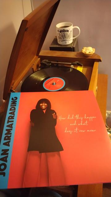 Record player with a 12" black vinyl record. In the foreground is an album cover showing Joan Armatrading against a red background, wearing a black jacket and red trousers. Behind the player is a mug of coffee on a speaker. There is also a mince pie on the corner of the player.