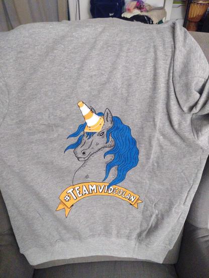 Sweat gris avec au dos une licorne à cheveux bleus. La corne étant un cône de chantier VLC. Et avec #TeamVideolan écrit.