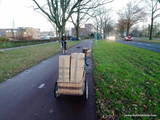 Bicycle pulling a trailer with some larger parcels of bicycle parts on a cycle-path on the way to the post office. You can also see the road with some cars and other cyclists on the cycle-path.