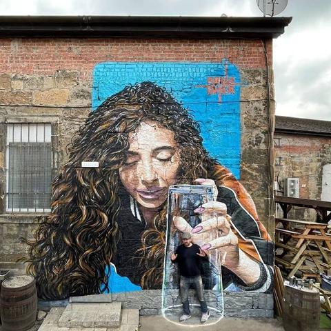 A mural by Bobby Rogue-One in Glasgow, Scotland, located at The Viceroy Bar & Club. The artwork depicts a woman with long, flowing curly hair, her eyes gently closed in a moment of serene focus. She holds an oversized drinking glass with her hand, its transparent surface skillfully painted to create a hyper-realistic effect. The mural's vivid blue background contrasts with the textured stone wall, blending photorealism and artistic creativity. The artist, visible in the image, stands in front of the mural as though enclosed within the painted glass, adding a playful, interactive dimension to the piece.