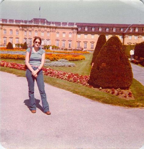 Young woman (16) standing on a walkway in front of a large building with ornate gardens in front of it.