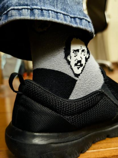 Acercamiento a un pie que lleva un tenis negro. El calcetín gris está decorado con la cara de Edgar Allan Poe.