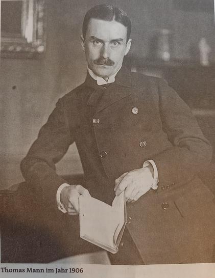 Ein Schwarzweiß Foto von Thomas Mann 1906. Erträgt einen Schnauzbart, schwarze  gegelte Haare mit Scheitel, einen dunklen Mantel mit zweireiigen Knöpfen und ein offenes Buch in der Hand. Mit einen Unterarm auf einem Möbelstück gelehnt.
Er hat sehr viel Ähnlichkeit mit C. M. Herbst. Nur in jünger und mit Haaren 😉