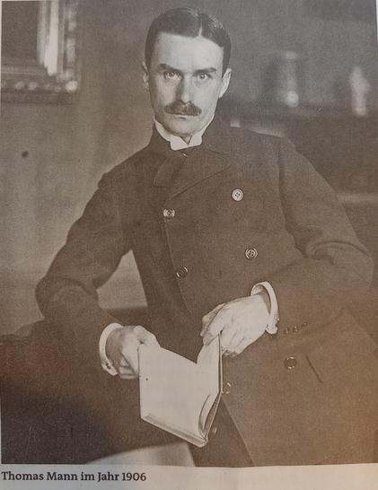 Ein Schwarzweiß Foto von Thomas Mann 1906. Erträgt einen Schnauzbart, schwarze  gegelte Haare mit Scheitel, einen dunklen Mantel mit zweireiigen Knöpfen und ein offenes Buch in der Hand. Mit einen Unterarm auf einem Möbelstück gelehnt.
Er hat sehr viel Ähnlichkeit mit C. M. Herbst. Nur in jünger und mit Haaren 😉