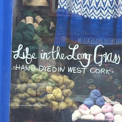 Außenansicht des Schaufensters von Bantry Yarns, ein Strickladen in Bantry, Irland. Stapel von Wollknäueln in Grüntönen und ein gestrickter Blau-weißer Pullover. In Spiegelschrift ist der Name des Pubs zu lesen, der auf der anderen Straßenseite liegt: Anchor Tavern.