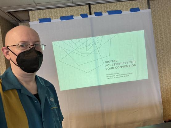 Me, a bald white man with glasses, wearing a black face mask and teal and mustard bowling-shirt style shirt (the Seattle Worldcon staff shirt, though the logo isn’t visible), in front of of an improvised projection screen made of a bedsheet blue-taped to a wall, showing an intro slide that says “Digital Accessibility for Your Convention”.