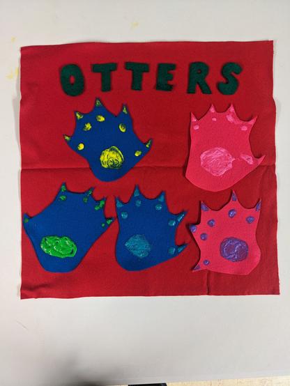 A red felt flag with the word "Otters" in green felt letters at the top. There are five otter paws made of felt arranged below the letters.