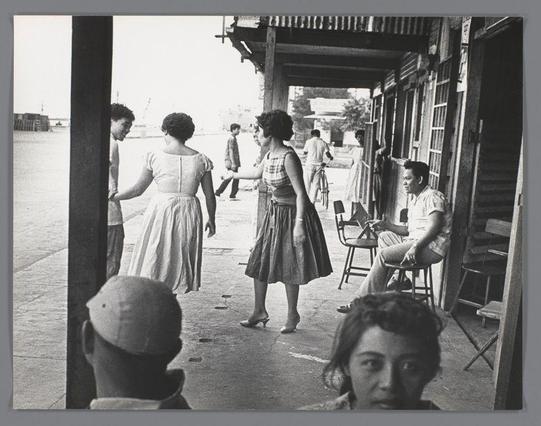 The image is a black and white photograph featuring several people in an outdoor urban setting. In the foreground, there's a man wearing a light-colored cap with his back turned to the camera, looking towards two women who are walking away from him on a paved street or sidewalk. One woman wears a sleeveless dress while reaching out for something or someone outside of the frame, and another follows her in a similar style but is more upright.

Behind them stands an older man seated casually with his legs crossed, holding what appears to be a drink container, possibly a soda bottle. He has dark hair styled upward and looks directly at the camera, exuding a relaxed demeanor. Other people can be seen around him: some are walking in various directions, while others sit or stand further back.

The architecture includes structures with exposed beams on the upper right side of the photo, suggesting an area that may have been industrial or repurposed for social gatherings such as bars and restaurants. The scene has a candid quality, capturing everyday interactions among people outside these establishments. 

This photograph is part of Ed van der Elsken's work documenting life in Cebu during 1959-60 at the Blue Dahlia Bar and Restaurant (formerly known as Susie's Blue), which provides insight into mid-century social scenes through his lens.

The image does not contain any visible text or context that would indicate a specific story [...]