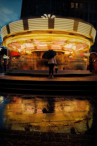 The image shows a brightly lit carousel at night, with blurred motion as it spins. A person with an umbrella is standing in front of it, and the ground is wet, reflecting the lights of the carousel. The scene has a vibrant, dynamic feel due to the motion blur and colorful lights.