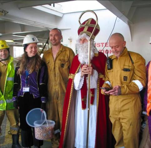 Ein als Bischof Nikolaus verkleideter Mann steht zwischen zwei Seemänner und neben den freiwilligen, die Helme mit dem Logo der Seemannsmission tragen