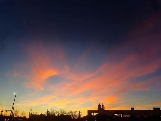 A stunning sunset sky with vibrant hues of orange, pink, and yellow blending into deep blue. Wispy clouds stretch across the sky, illuminated by the warm colors of the setting sun. Silhouettes of trees, streetlights, and a building outline the foreground, adding contrast and depth to the serene and picturesque scene.