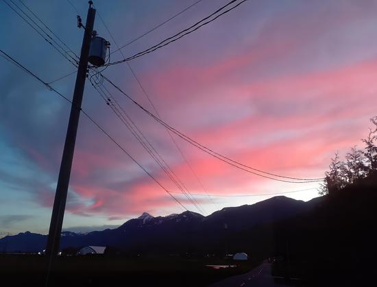 A breathtaking twilight scene with a sky painted in soft shades of pink, purple, and blue. The foreground features a utility pole with a web of power lines cutting across the sky, adding an industrial touch to the natural beauty. In the distance, rugged mountain peaks capped with patches of snow create a dramatic silhouette. A narrow road winds through the scene, flanked by trees and open fields, while faint structures and lights hint at human habitation. The vibrant clouds and serene landscape combine to evoke a sense of peaceful evening tranquility.
