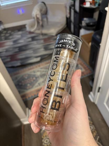 Photo of a white person’s hand holding a clear tube with 5 small containers of honey snacks. The brand is Jamie’s Hive To Table and the product is Honeycomb Bites. In the background is a white greyhound dog with a black spot preparing to curl up in her bed.