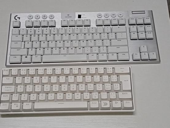 Foto mostrando dois teclados sobre uma mesa. O de cima é um Logitech G915 TKL, que possui uma base de metal escovado e teclas brancas; o de baixo é um KBM! TG600 (60%), a princípio, suas teclas são brancas, mas mesmo novas elas possuem um tom "amarelado".