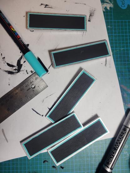 Wooden rectangles painted black with silver trim and light teal interior highlights sit on a white piece of paper with a marker and ruler.