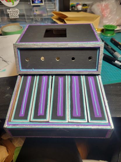 Front view.
A homemade keyboard /organ painted black with silver trim, teal highlits on the corners and edges, and purple interior trim. The keys now have a teal line through a purple rectangle down the middle length of the keys