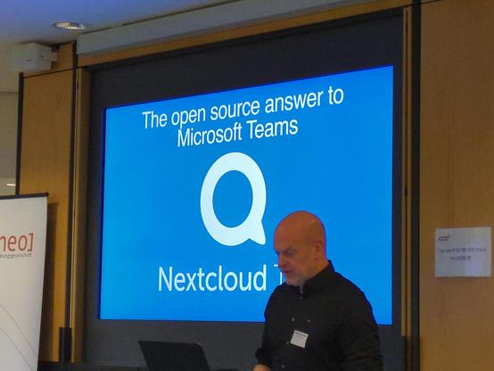 Frank Karlitschek hält eine Präsentation über Nextcloud und vergleicht mit Microsoft Teams. Im Hintergrund ist die Präsentation mit blauem Hintergrund und weißer Schrift zu sehen, im Vordergrund Frank Karlitschek.

Der Text auf der Präsentation sagt:

The open source answer to Microsoft Teams

- dann folgt dann Nextcloud Logo groß in der Mitte - 

Nextcloud Talk