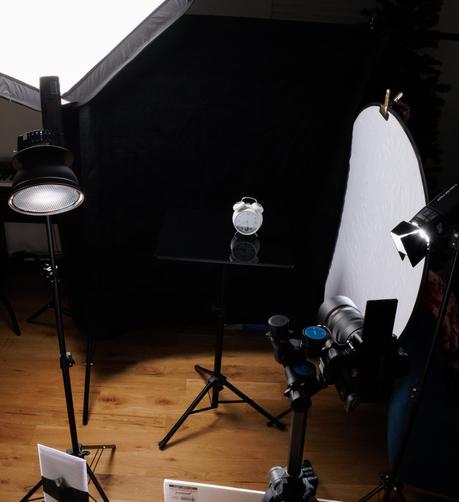 A behind-the-scenes setup of a tabletop photography session with a white alarm clock on a reflective black surface. The scene is surrounded by various studio equipment, including softbox lights, a reflector, and a camera on a tripod. A black fabric backdrop isolates the subject.