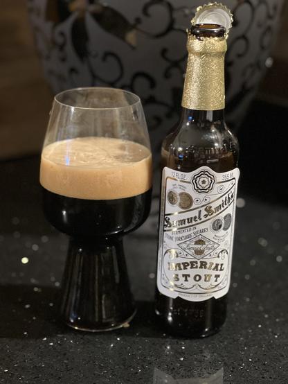A stout glass full of dark beer with a thick tan head. The brown bottle has a gold foil neck label and a white label with scripture text in shades of gold and black