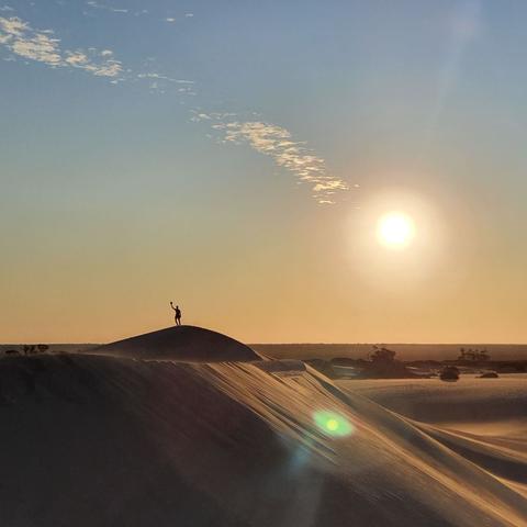A person stands silhouetted atop a sand dune during sunset. The sun is low in the sky, casting a warm golden glow across the scene.