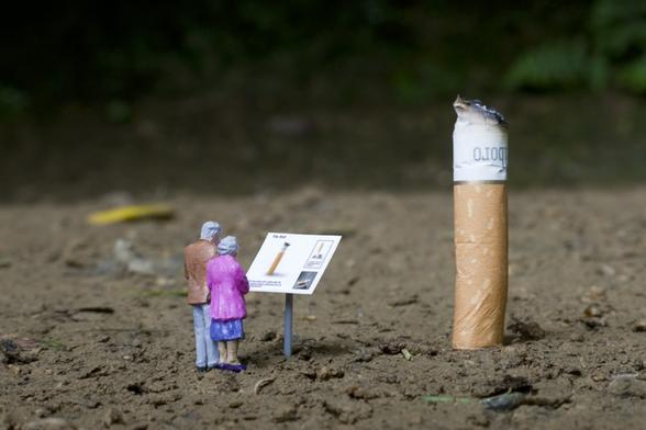 Artwork from the 'Little People' series by Slinkachu, featuring two miniature figurines, an elderly man and woman, standing in front of a sign resembling a museum exhibit description. The sign explains a cigarette butt, which looms large in comparison to the tiny figures, turning the discarded object into an oversized, thought-provoking display. The scene is set on a patch of dirt, blending urban commentary with whimsical scale manipulation.