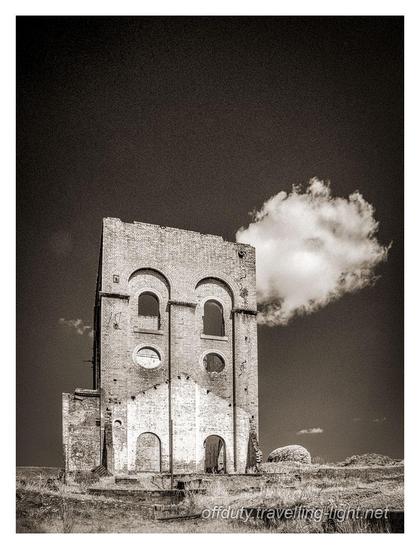Sepia toned infrared black and white photo of the shell of an old industrial building against a dark sky, with a single cloud partly hidden.