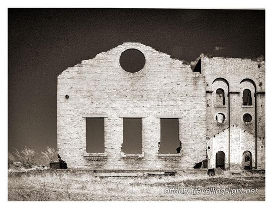 Sepia toned infrared black and white photo of the shell of an old industrial building against a dark sky. Thre is a wall with three windows at ground floor level and a circular hole high in the gable end.