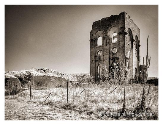 Sepia toned infrared black and white photo of the shell of an old industrial building against a dark sky, with overgrown low ruins around.
