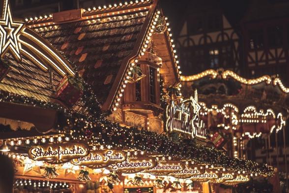 Eine stimmungsvolle Aufnahme eines traditionellen deutschen Weihnachtsmarktstandes bei Nacht. Der Stand ist im typischen Holzhütten-Stil gebaut, mit einem spitzen Dach und ist festlich geschmückt. Die gesamte Struktur ist mit warmweißen Lichterketten umrandet, die einen goldenen Schein erzeugen. An der Front sind beleuchtete Schilder mit deutscher Beschriftung zu sehen, darunter "Schokofrüchte" und "Erdbeeren". Dekorative goldene Sterne und Weihnachtsglocken mit Tannengrün schmücken die Dachkante. Im Hintergrund sind weitere beleuchtete Marktstände zu erkennen, die eine typische Weihnachtsmarkt-Atmosphäre schaffen. Die warme Beleuchtung erzeugt eine gemütliche, festliche Stimmung, die charakteristisch für deutsche Weihnachtsmärkte ist.