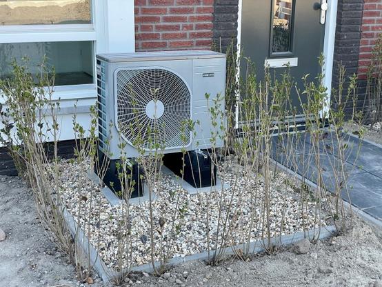 Außeneinheit einer Luft-Wärmepumpe, montiert an einer roten Ziegelwand eines Gebäudes. Die Wärmepumpe ist in hellgrauer Farbe ausgeführt und verfügt über einen großen, kreisförmigen Ventilator mit Schutzgitter. Das Gerät ist auf einem erhöhten Betonsockel installiert, der mit weißem Kies und jungen Sträuchern umgeben ist. Im Hintergrund sind weiße Fensterrahmen und eine graue Tür zu erkennen. Die Bepflanzung um die Wärmepumpe herum zeigt erste Frühjahrstriebe mit kleinen grünen Blättern.