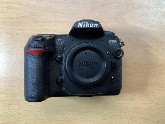 Cámara Nikon D200. Es negra con logos en blanco de Nikon y D200. Tiene una línea roja característica de Nikon en la empuñadura, donde apoyas la mano derecha, debajo del disparador.
