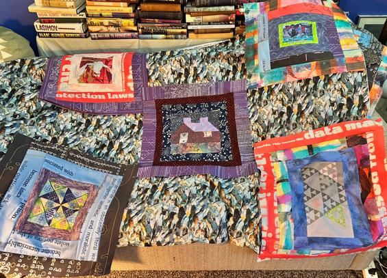 This image shows a collection of vibrant quilt blocks, each featuring intricate patterns and varied themes. The designs integrate elements of patchwork and printed text, with vivid colors and abstract imagery. The blocks rest on a quilted background patterned with textured imagery resembling natural elements. A bookshelf in the background holds a variety of books.