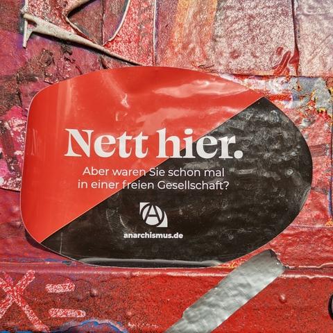 Here's alt text for the image:A sticker, half red and half black, is affixed to a textured, red surface. The sticker features the German words "Nett hier," meaning "Nice here," in bold white font. Below it, smaller text poses the question, "But have you ever been in a free society?" A logo with the letter "A" inside a circle and the website anarchismus.de are also visible at the bottom. The background is a heavily painted or textured surface with layers of red and other colors.