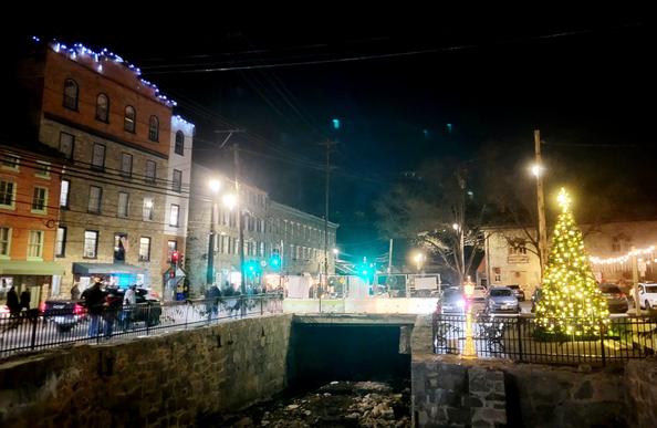 Night photo,  lighted large fake Christmas tree on the right bank of a culvert with a road bridge. Left bank is historic main street shops and shoppers. Behind the tree is the railroad museum.