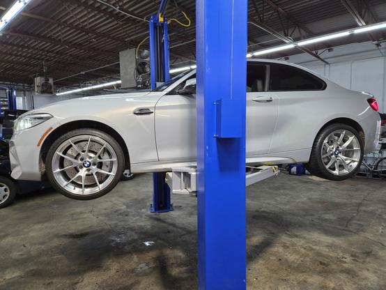 A light grey BMW M2 on a blue automotive lift in a shop.