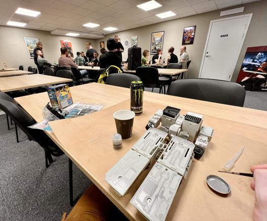 G1 Metroplex being panel lined in the foreground, Warhammer league gathering in the background, lots of hobby tables on the room and fun geeky posters on the wall