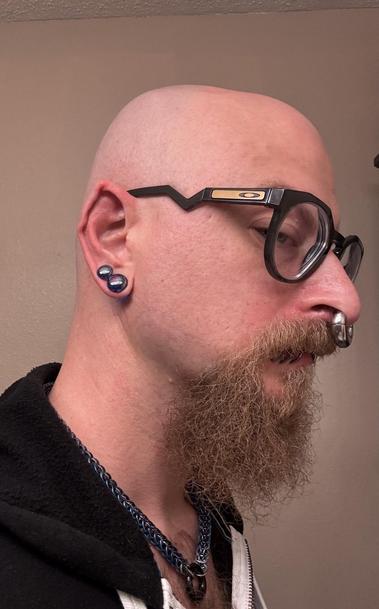 A bald person with a beard is shown in profile. They wear large, distinctive glasses and have multiple facial piercings, including a septum ring and lip piercings. Their ears are pointed with double plugs in their earlobes are visible, and they have a blue chain around their neck.