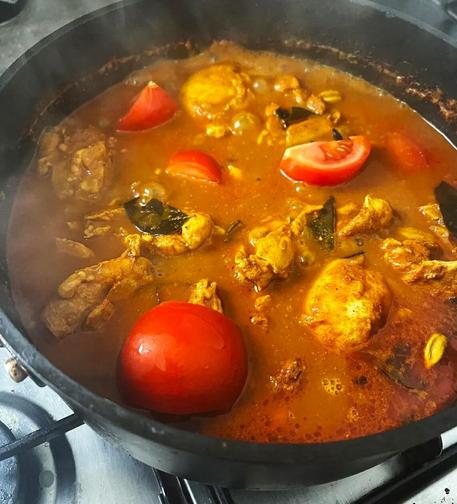 A brown-orange curry broth with orange chicken, bright red tomatoes and black curry leaves in a black steel pan