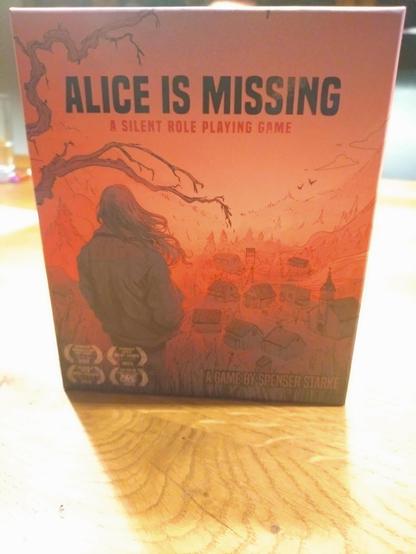 Eine in rot gehaltene Spielpackung. In grau und schwarz Tönen ist in der Ferne ein Dorf und im Vordergrund die Rückseite eines Mädchens zu sehen.
Auf der Box steht: Alice is missing
A silent Role Playing Game