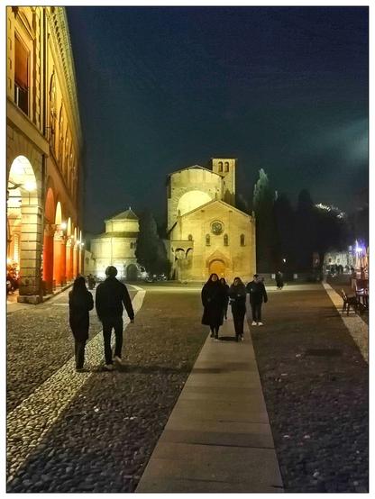 Una vista notturna di una plaza, con una coppia che si allontana dalla macchina fotografica verso una grande chiesa illuminata. Anche altre persone camminano nella piazza. A sinistra si accende anche un lungo edificio ad arco.