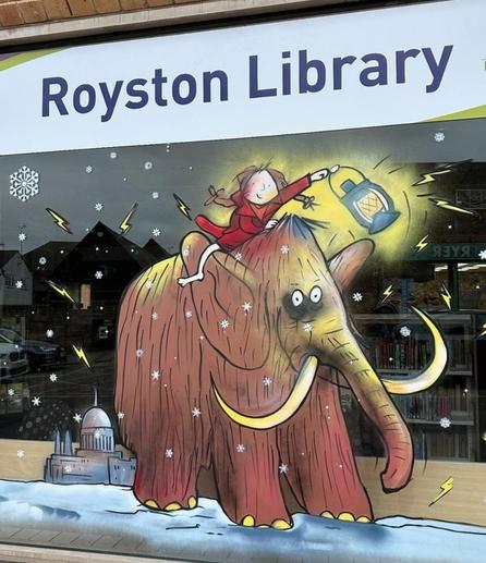 Streetart. A cute, Christmassy picture of a mastodon was painted on the window of a library. It shows a little brown-haired girl in red pyjamas holding a glowing lantern and riding a shaggy brown mammoth through the snow. A cathedral and stars can be seen in the background. A very cute idea for a library. 
Info: The original drawing was created by Quentin Blake for the book "The Ice Monster" by British comedian and author David Walliams. Sir Quentin Saxby Blake CH (born December 16, 1932) is a fantastic British cartoonist, illustrator and children's book author.
