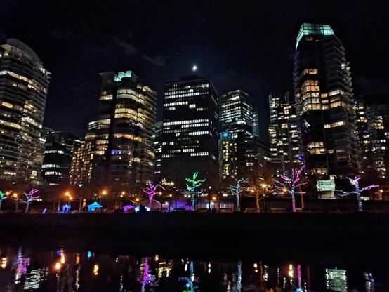 El paseo marítimo de Vancouver de noche, con luces navideñas, visto desde el mar: una hilera de arbolitos de colores que se reflejan en el agua y, tras ellos, rascacielos iluminados.
